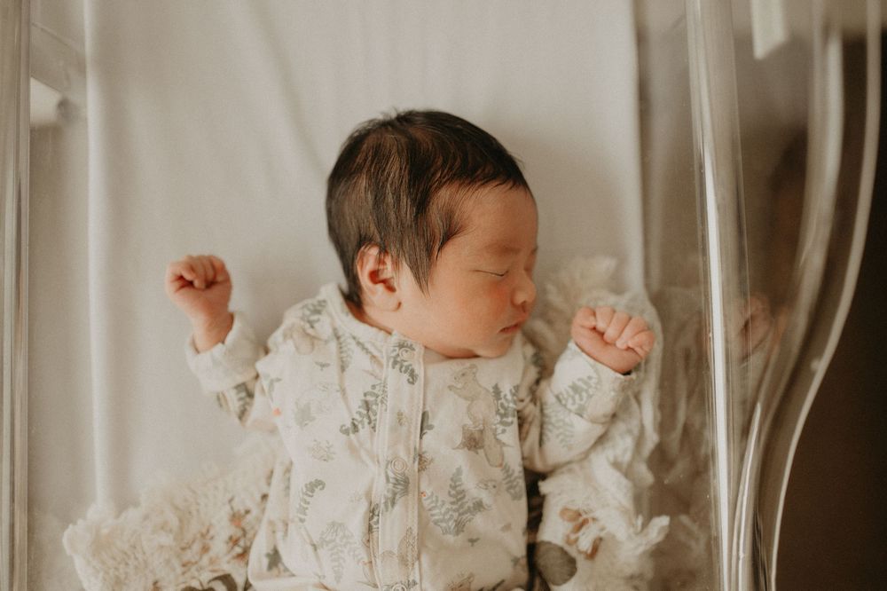 Sleeping newborn baby in a floral print outfit lying in a hospital bassinet with tiny hands raised near the head.