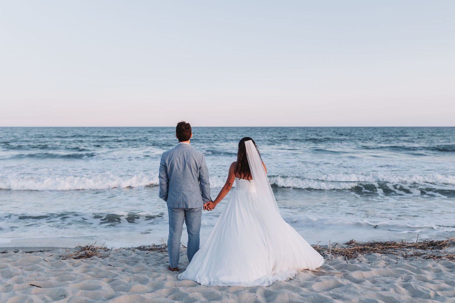 Bride in a lace wedding gown with a flowing veil holds a blue bouquet on a rocky beach while the groom in a navy suit stands nearby.