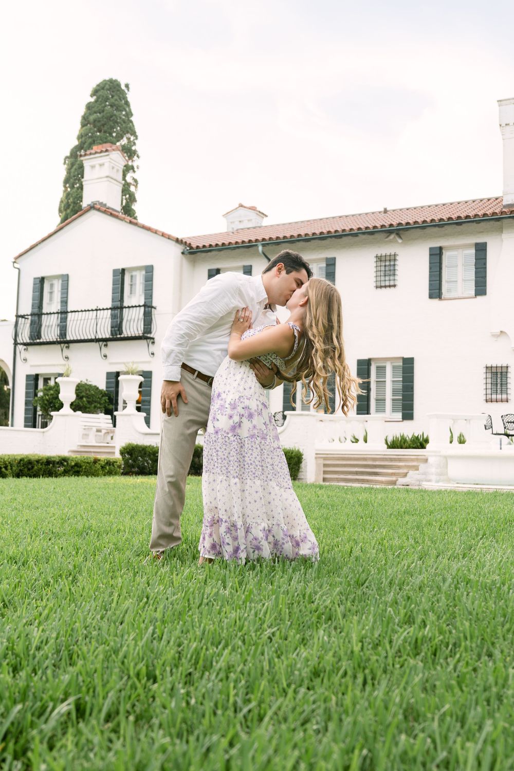 Wide sweeping shot of couple kissing on the lawn in front of Crane Cottage during their Jekyll Island engagement photography session.