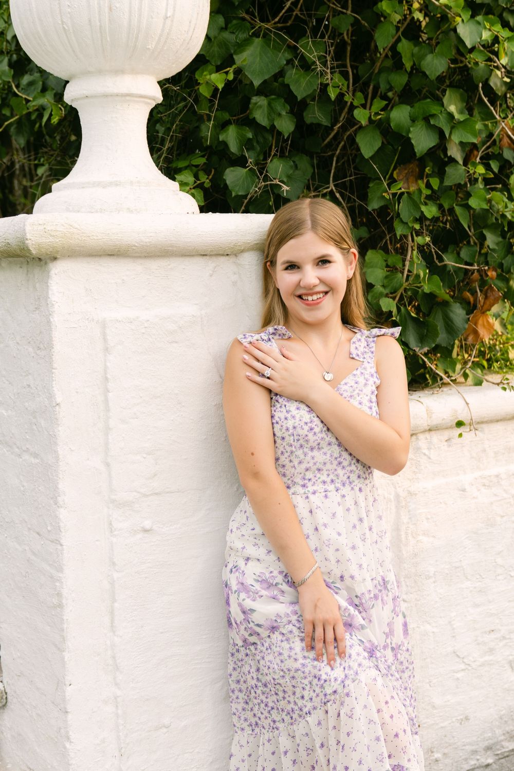 Woman in a lavender floral dress showing her engagement ring beside a white garden wall and urn on Jekyll Island after just getting engaged