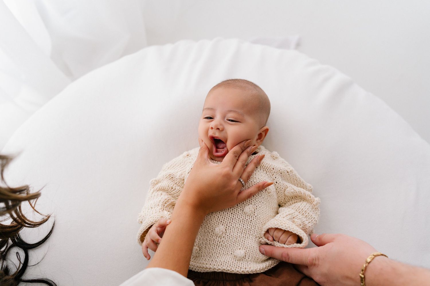 A laughing baby in a cream knit sweater being held by a mother in a white round chair.