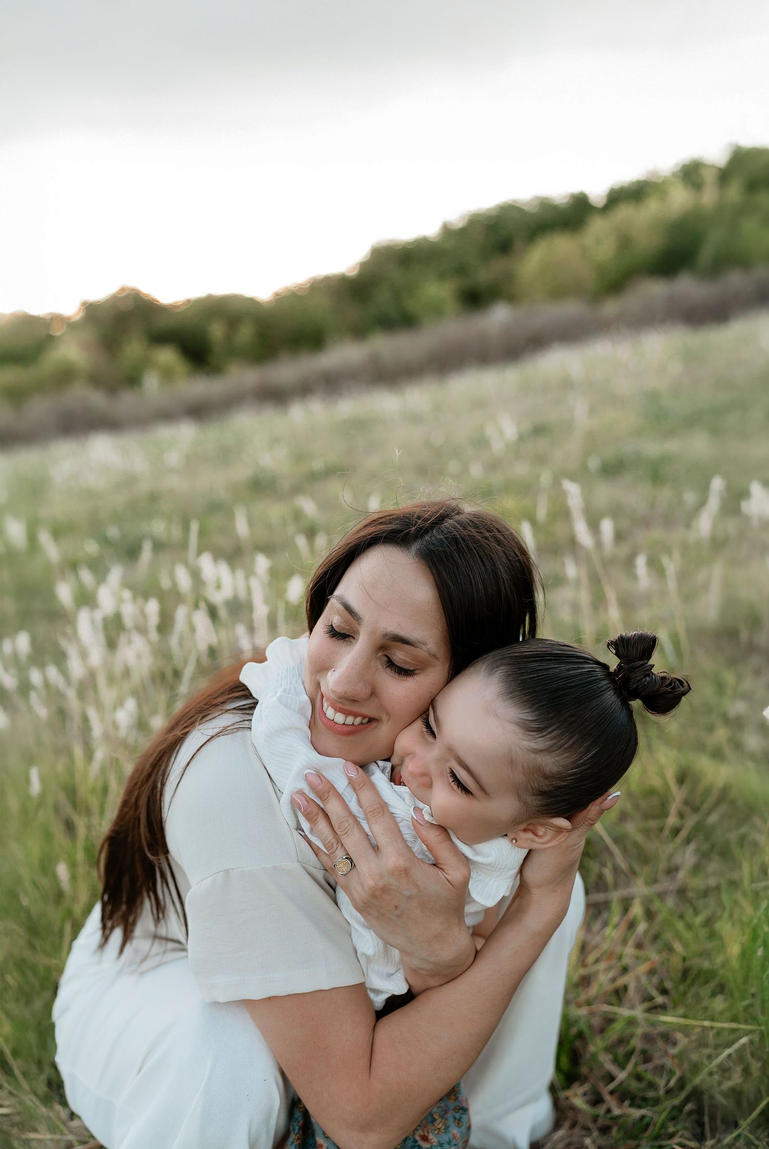 A mother lovingly embraces her young daughter in a sunlit meadow, both wearing white in a tender outdoor moment.