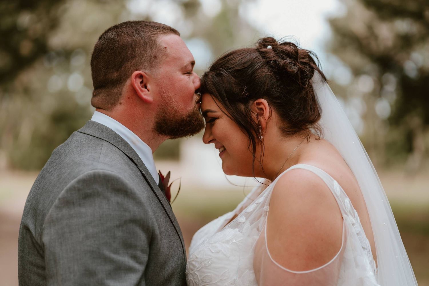 A groom in a gray suit kisses his bride on the forehead as she smiles in a white lace dress and veil.