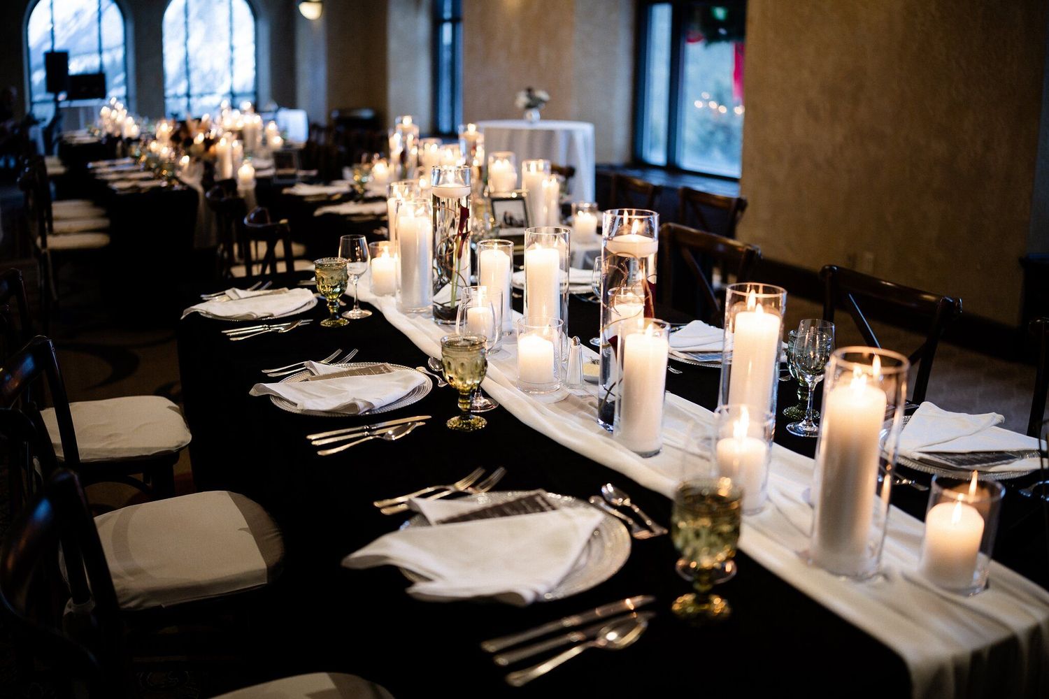 Elegant wedding reception table with black linen, white pillar candles, and formal place settings in a dimly lit venue.