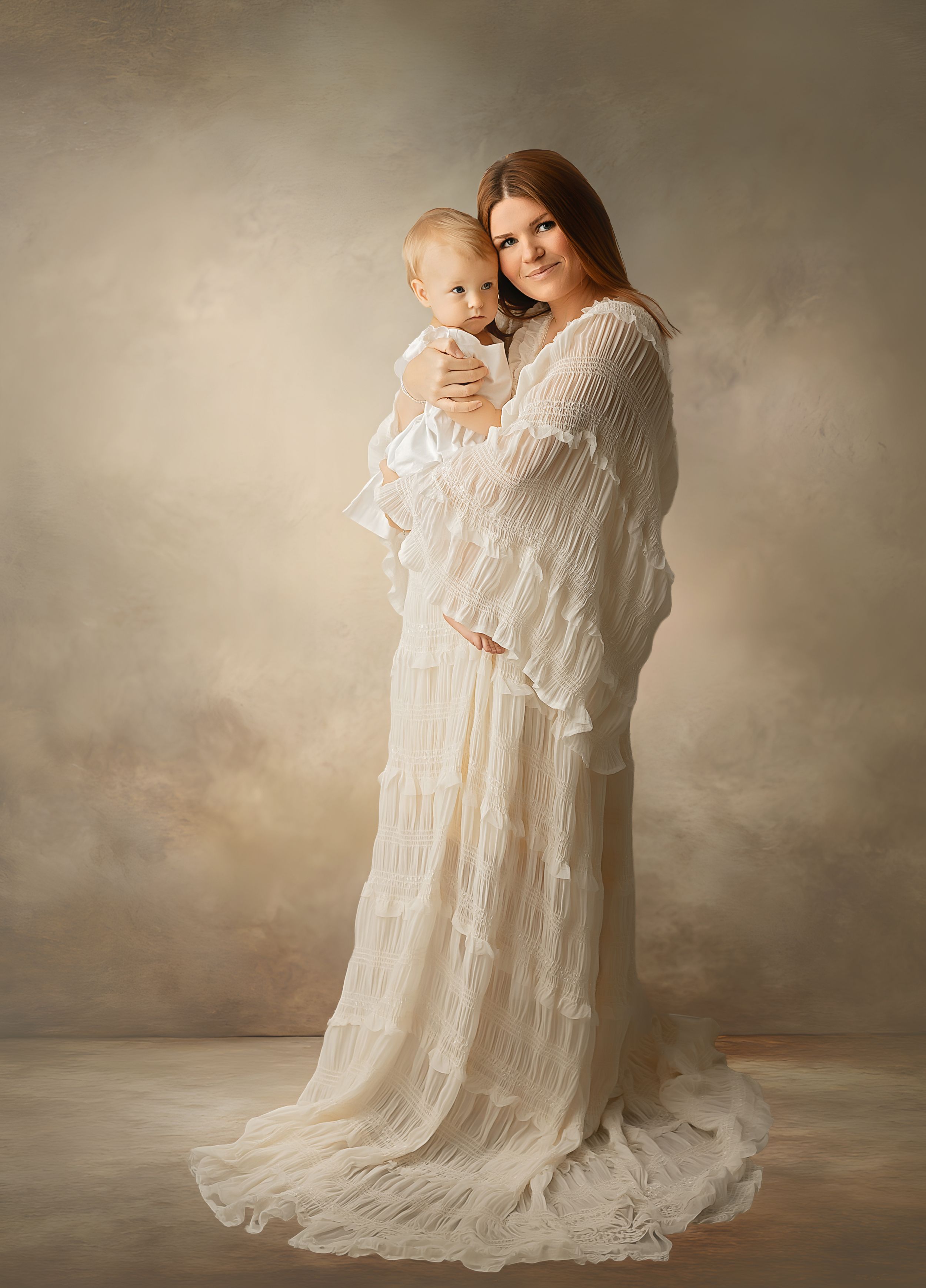 A mother in a flowing white gown holds her baby during an elegant studio portrait session.