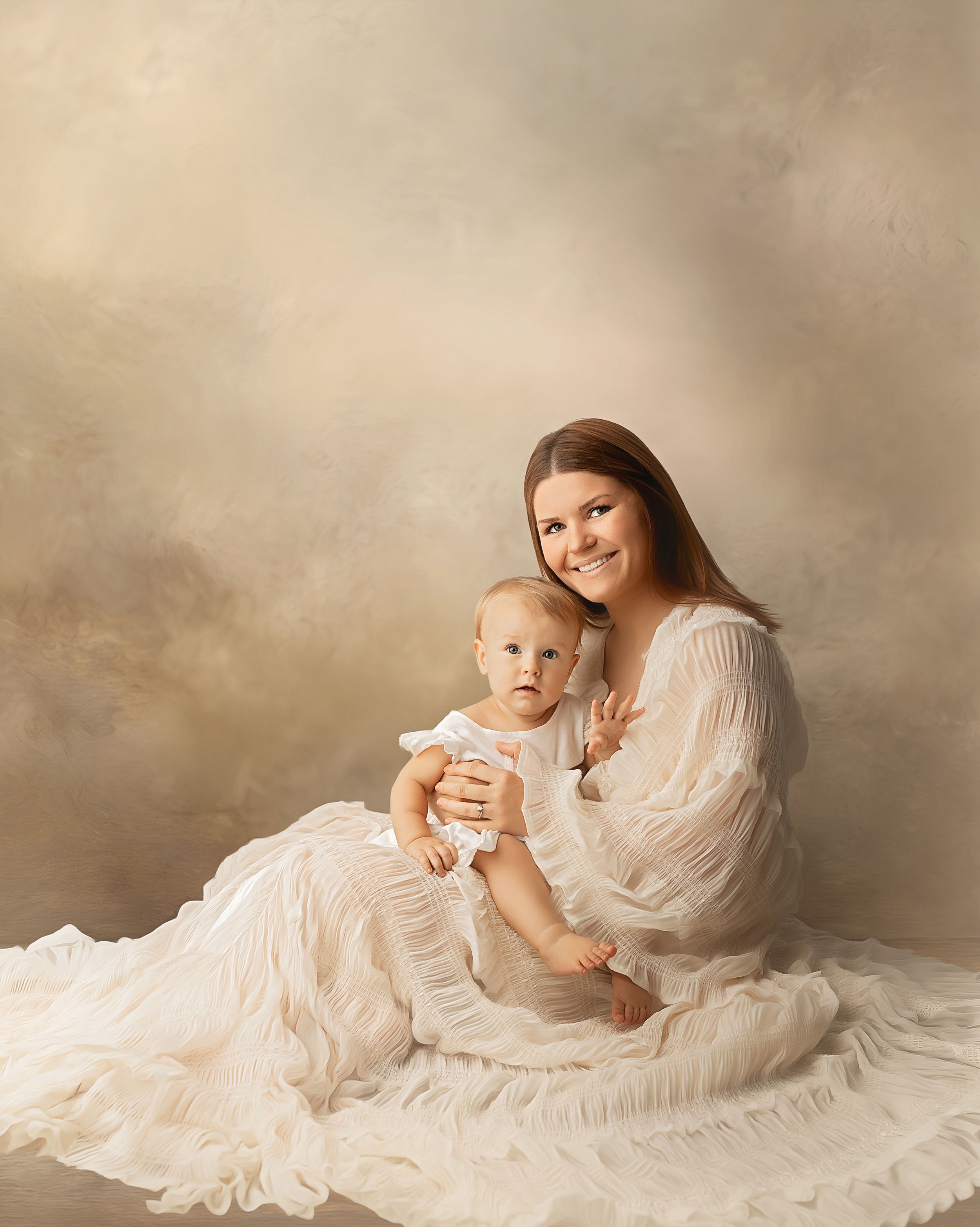 A smiling woman in a white dress sits with a baby in a soft, elegant studio portrait with neutral tones.