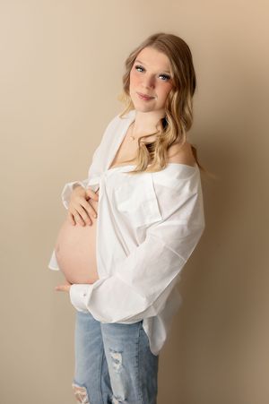 Pregnant woman in white off-shoulder shirt and ripped jeans cradling baby bump in a studio portrait.