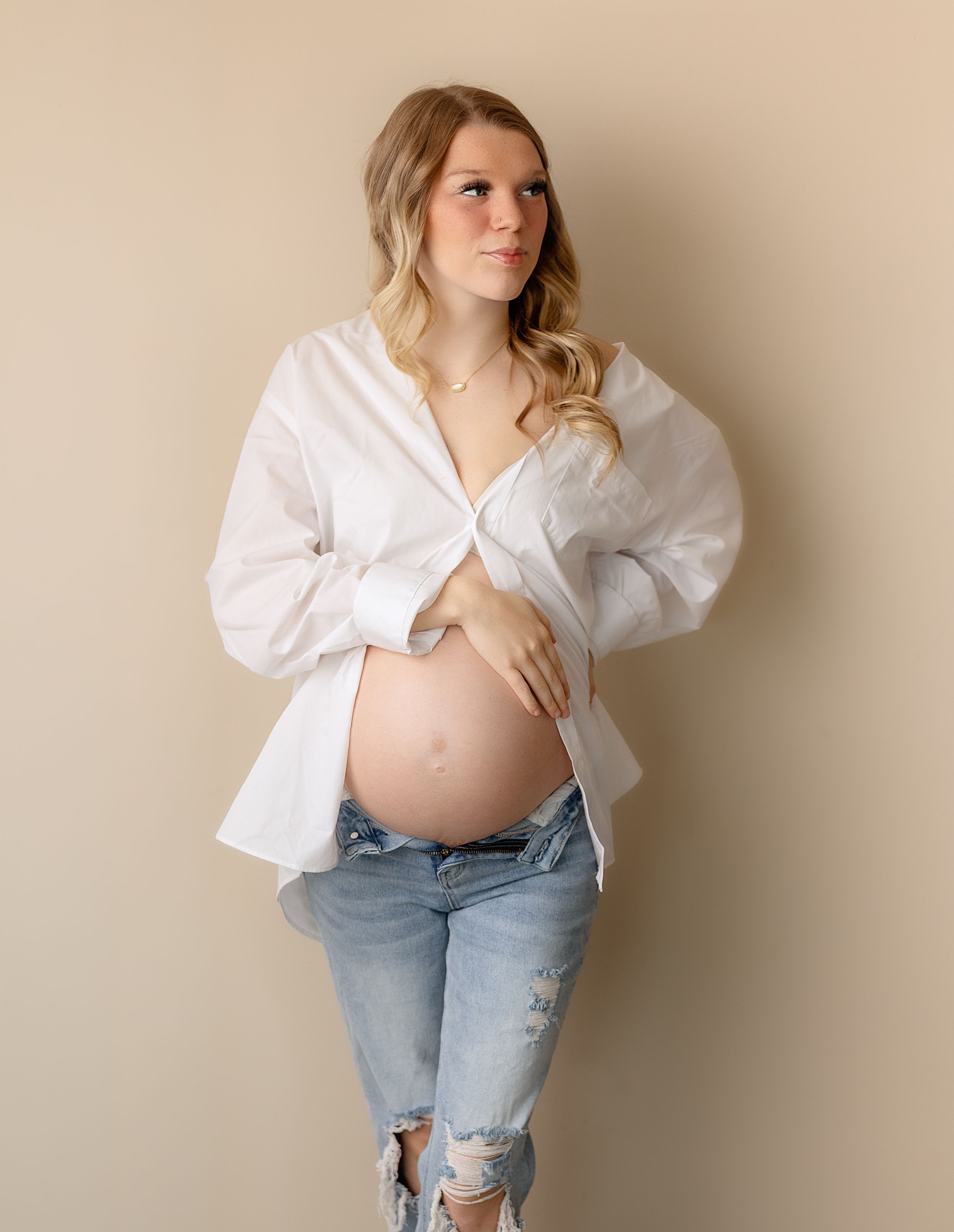 Pregnant woman in white open shirt and ripped jeans cradling her baby bump against a neutral beige background.