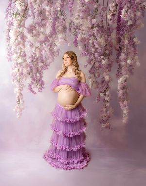 Pregnant woman in purple tulle gown poses among hanging wisteria flowers in elegant maternity portrait.