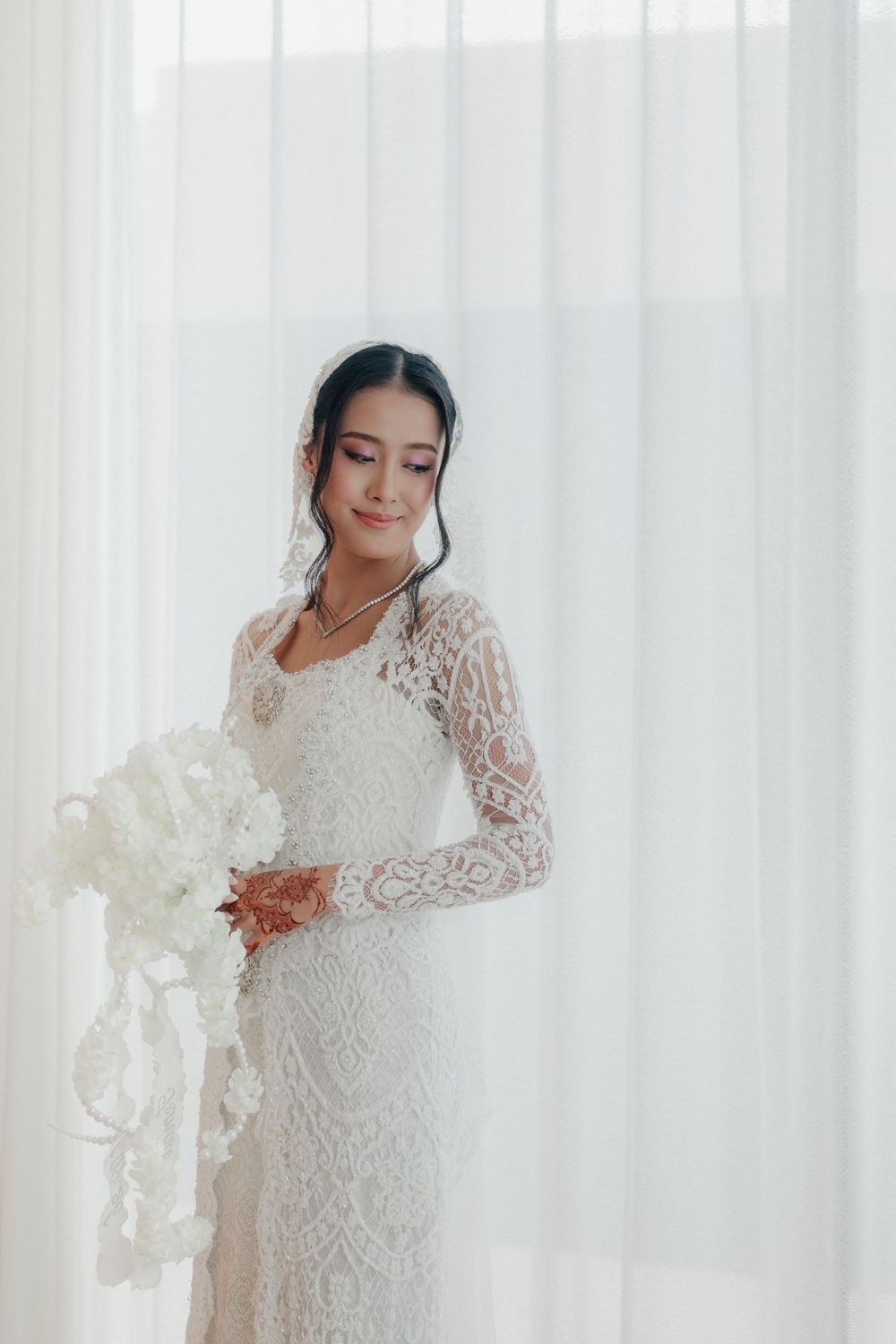 Elegant bride in long-sleeve lace wedding dress holding white floral bouquet, standing by sheer curtains in soft natural light.