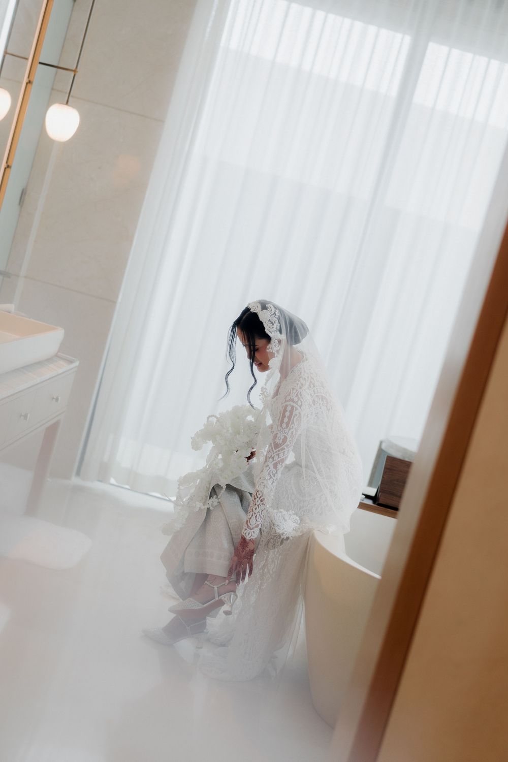 Bride in white lace wedding dress and veil holds bouquet by bright window in elegant bathroom.