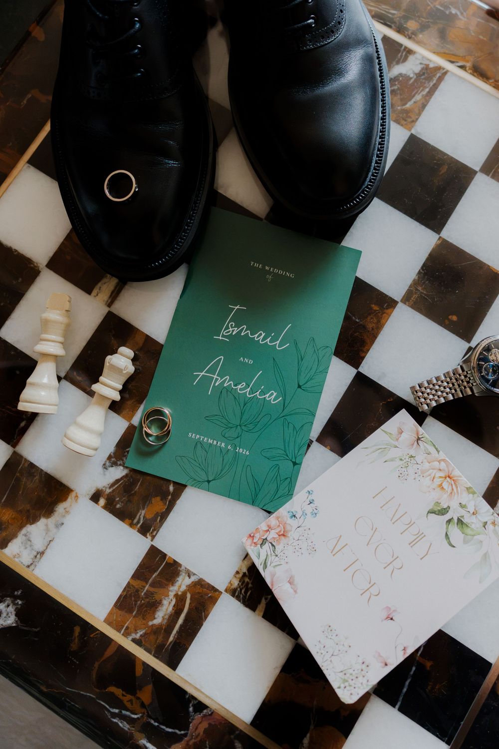 Wedding flat lay featuring green invitation for Ismail and Amelia, rings, watch, chess pieces, and black dress shoes on marble board.