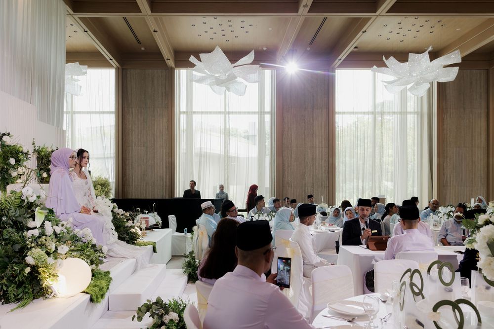 Elegant wedding reception hall with white floral decor, guests in formal attire seated at tables, and a bride in a white hijab.