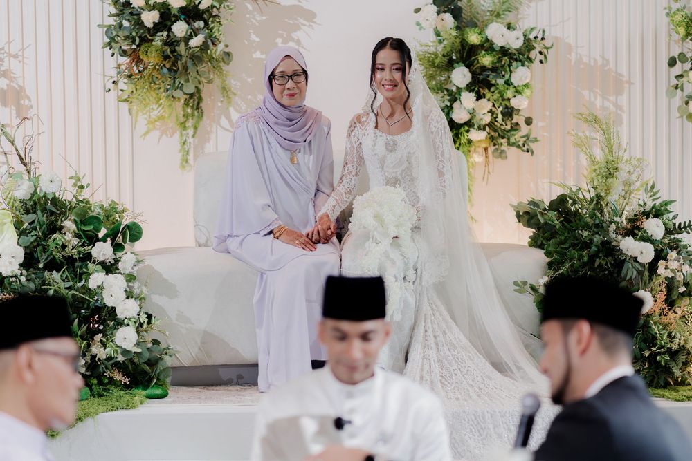 A bride in white lace gown poses with a woman in lilac hijab at an elegant floral wedding ceremony.
