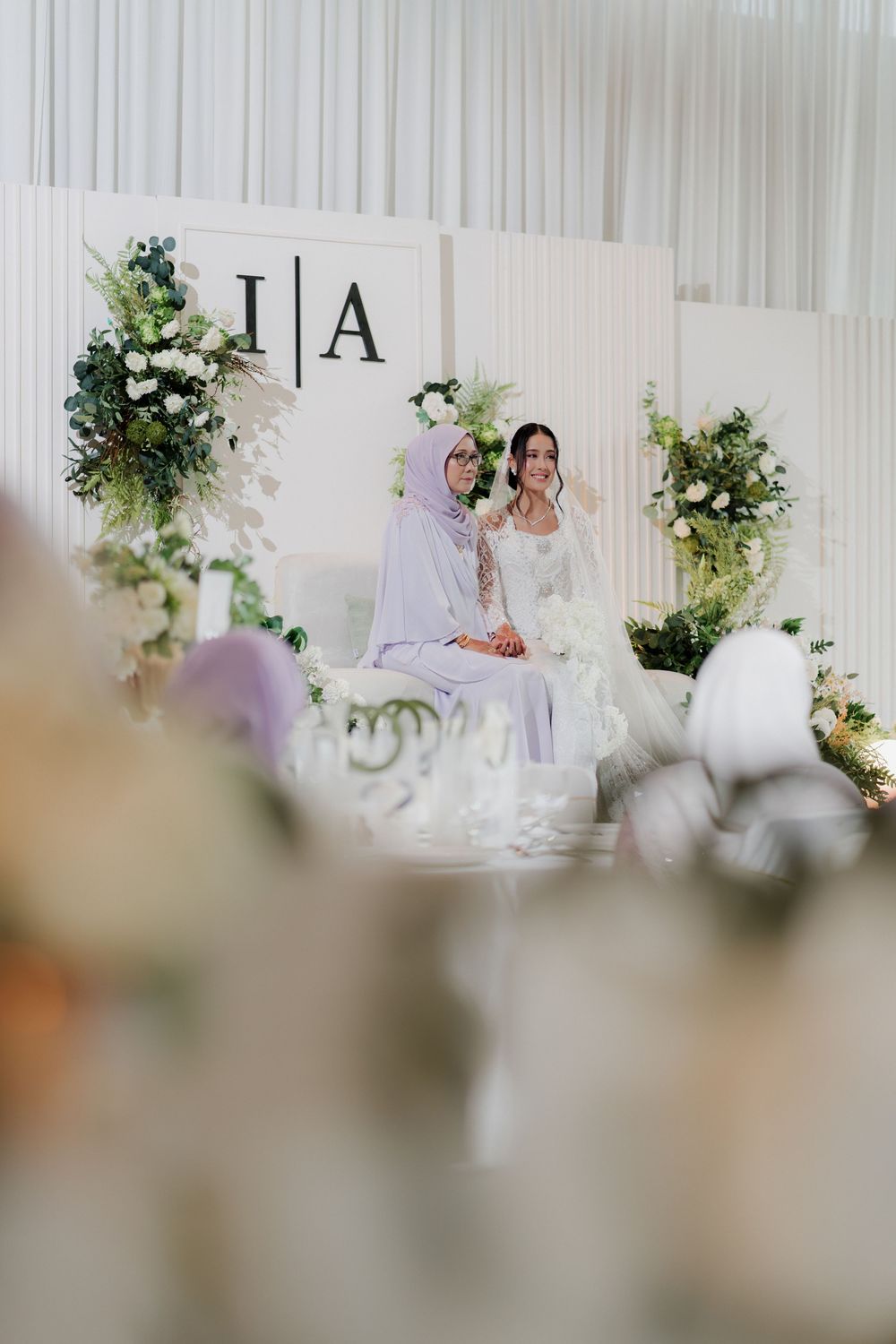 Two women in lavender attire pose at an elegant wedding reception backdrop with floral arrangements and monogram decor.