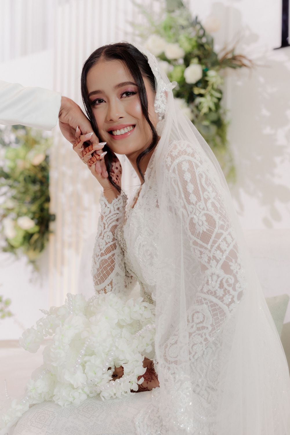 Radiant bride in white lace gown and veil holds bouquet, smiling beautifully at her wedding ceremony.