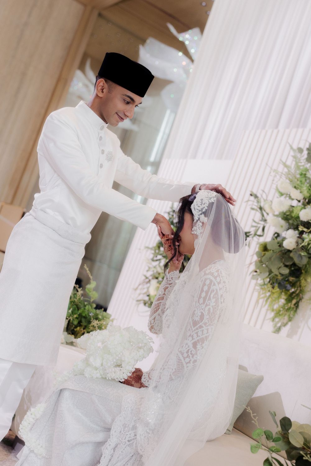 Malay groom in white baju melayu and songkok places veil on seated bride during traditional wedding ceremony.