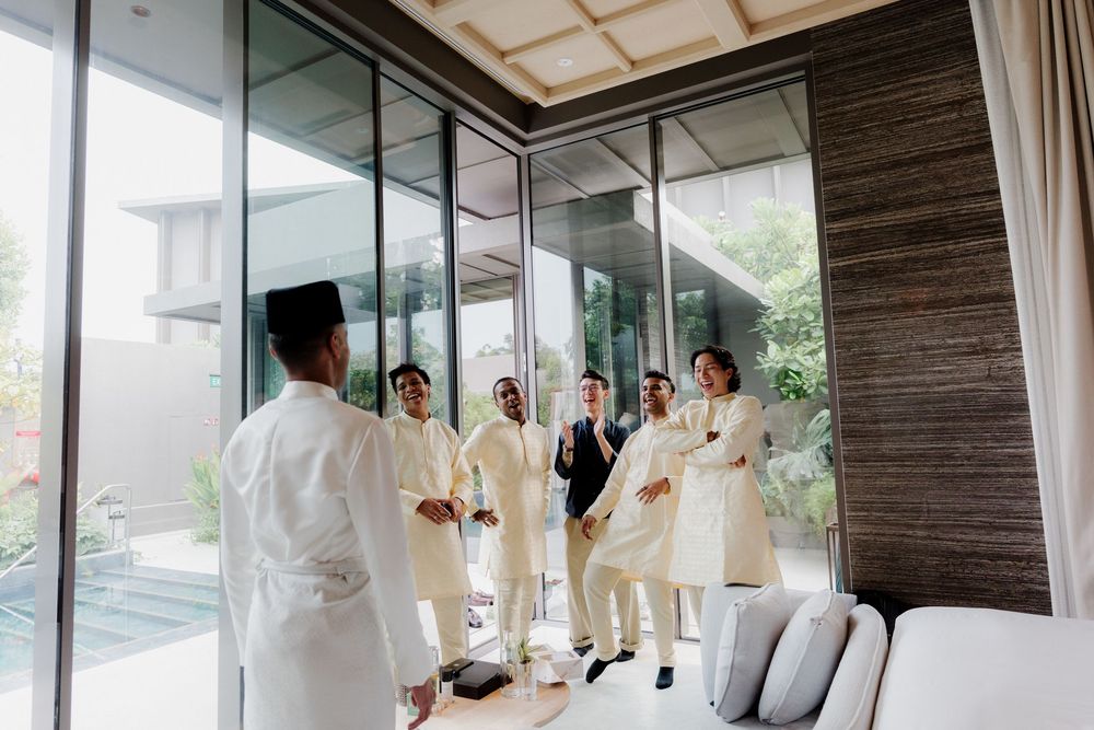 Groom and groomsmen in white traditional attire gather indoors before a wedding ceremony.