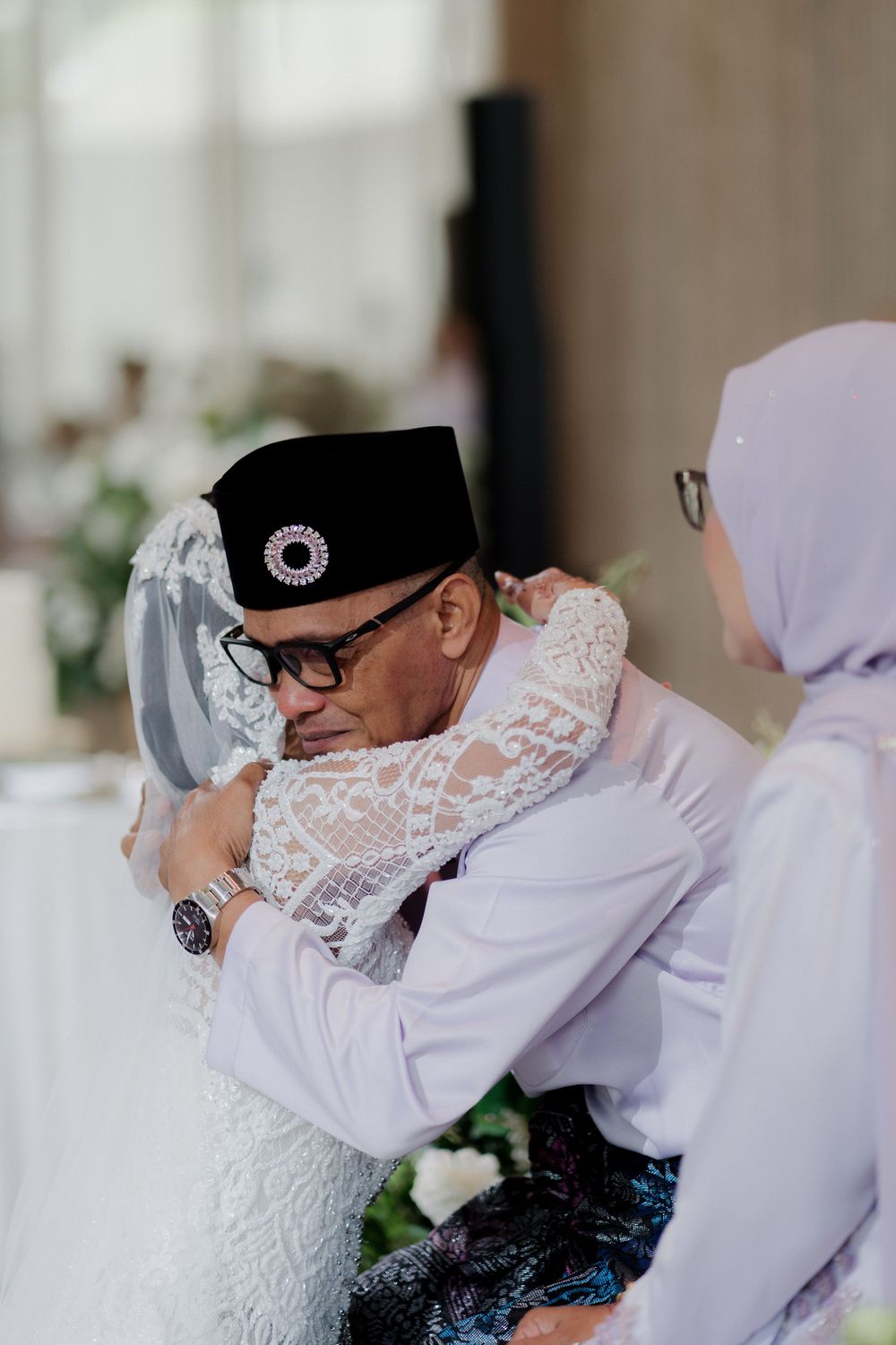 Emotional wedding moment as bride in white lace dress embraces older man wearing traditional black peci hat.