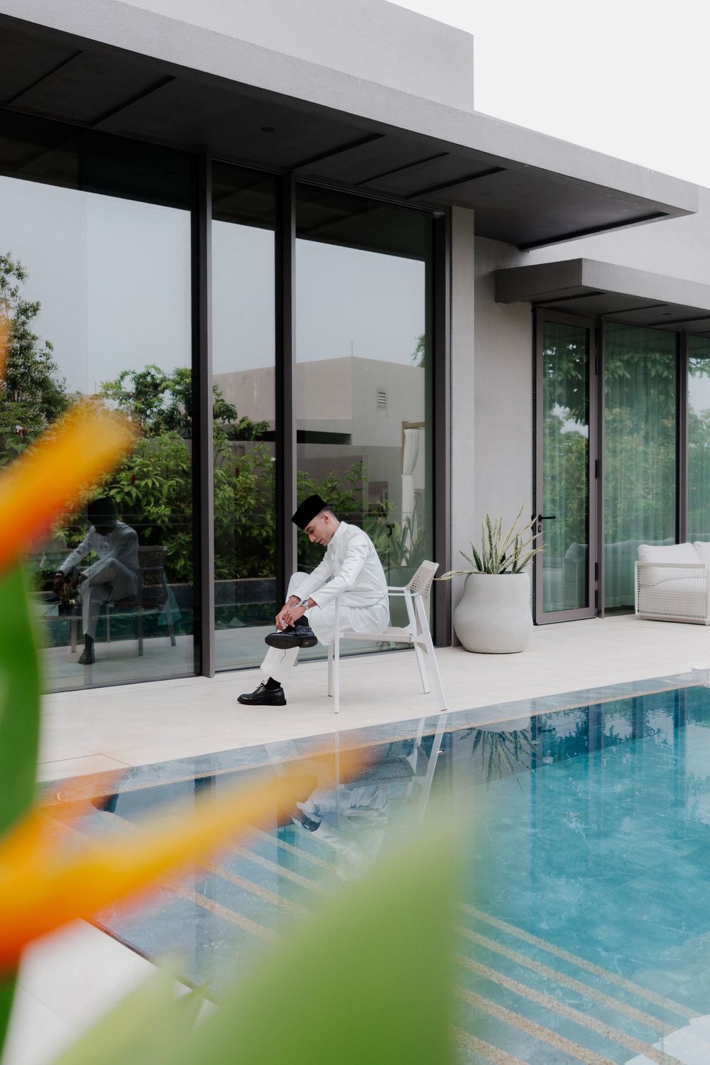 Man in white outfit sitting poolside at a modern luxury villa with large glass windows and tropical landscaping.