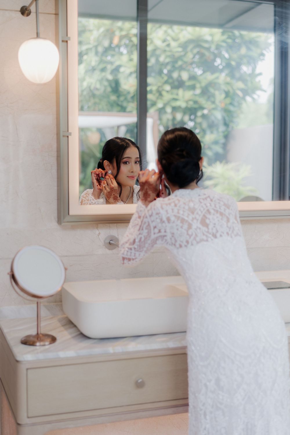 Bride in white lace dress applying makeup at bathroom vanity mirror on her wedding day.