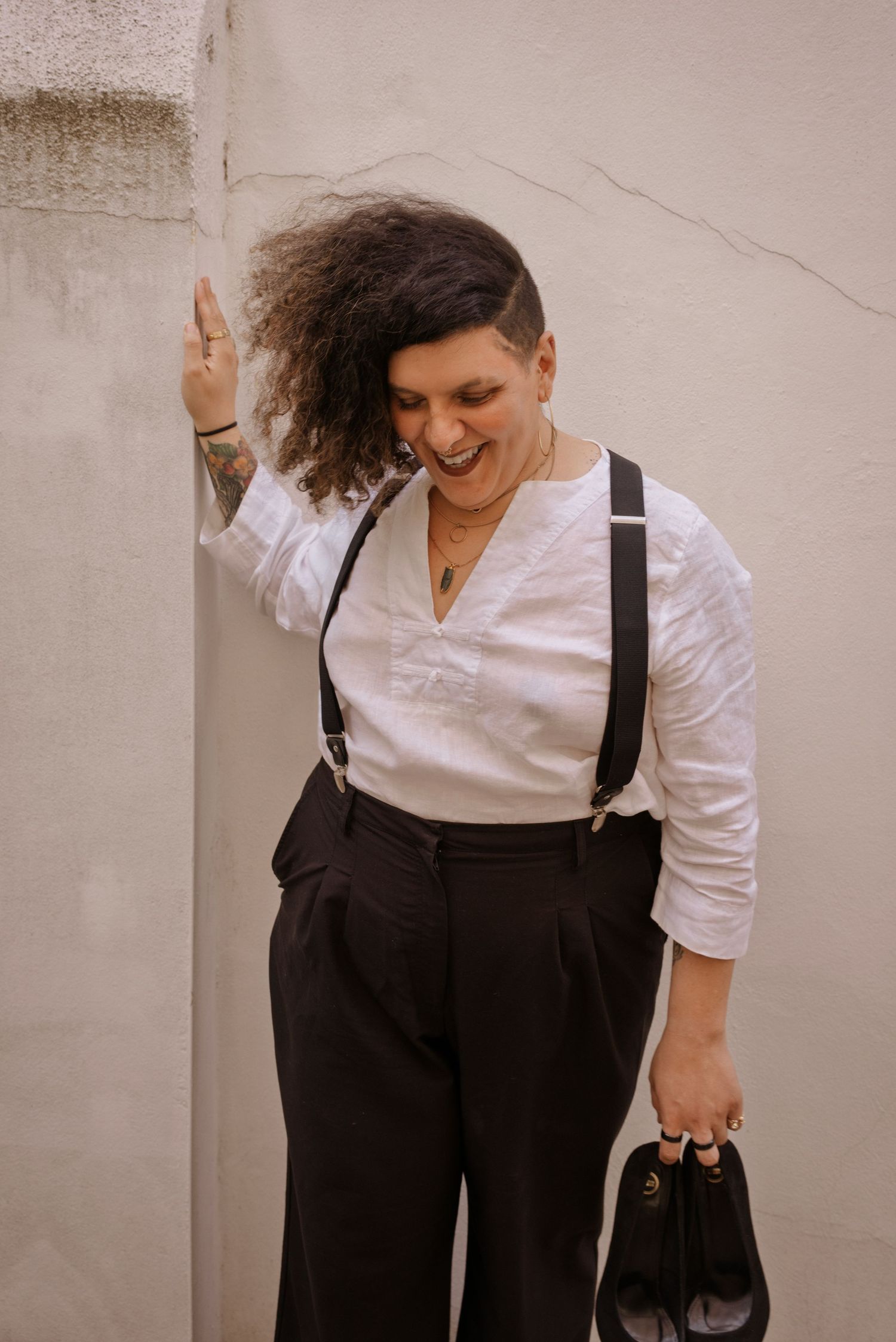 Person in white shirt and black suspenders with wide-leg trousers, laughing while leaning against a wall.