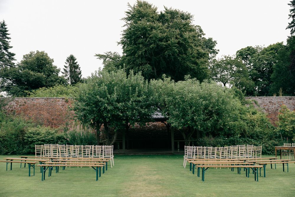 Outdoor wedding ceremony setup with wooden chairs on green lawn, surrounded by lush hedges and brick wall garden.