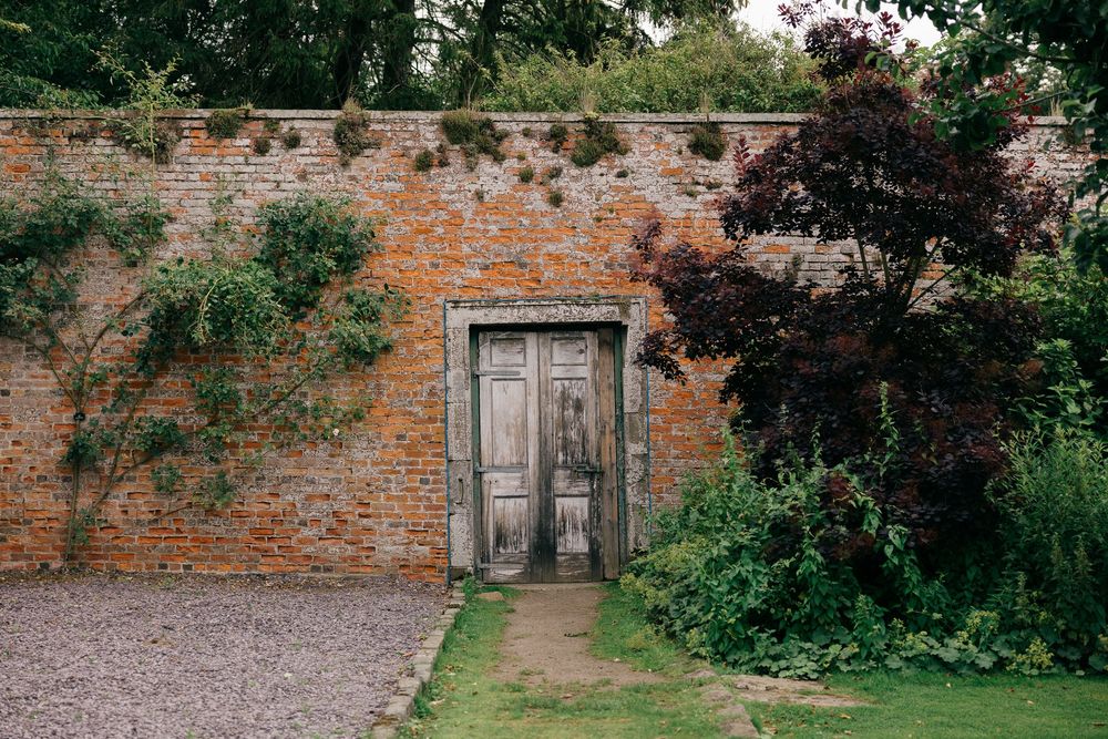 Rustic weathered wooden doors set in an old brick garden wall surrounded by lush greenery.