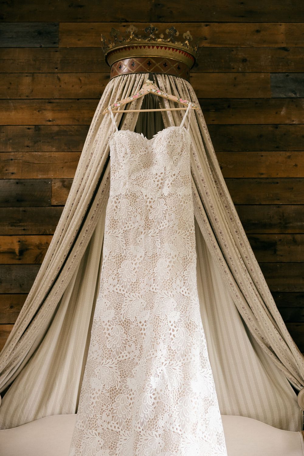 Elegant lace wedding dress hanging on a hanger beneath a decorative crown canopy against a wooden wall.