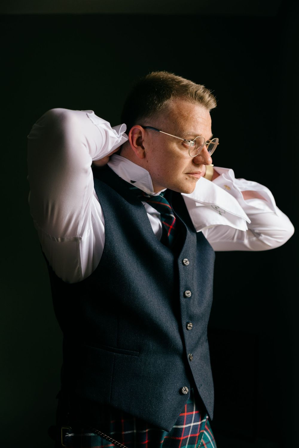 Groom in tartan kilt and navy waistcoat adjusting his collar against a dark background.