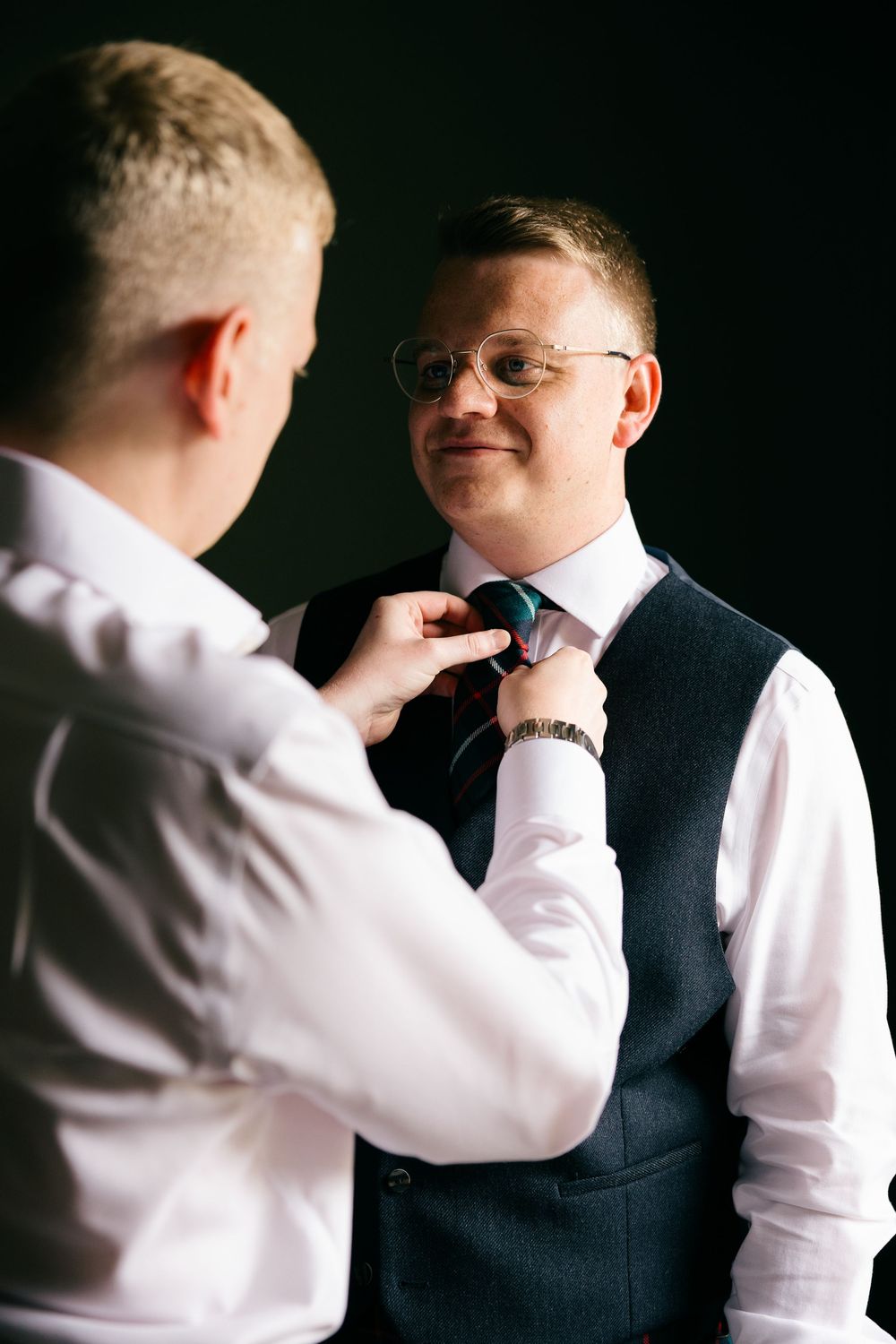 Groomsman adjusts groom's tie during wedding preparations indoors.