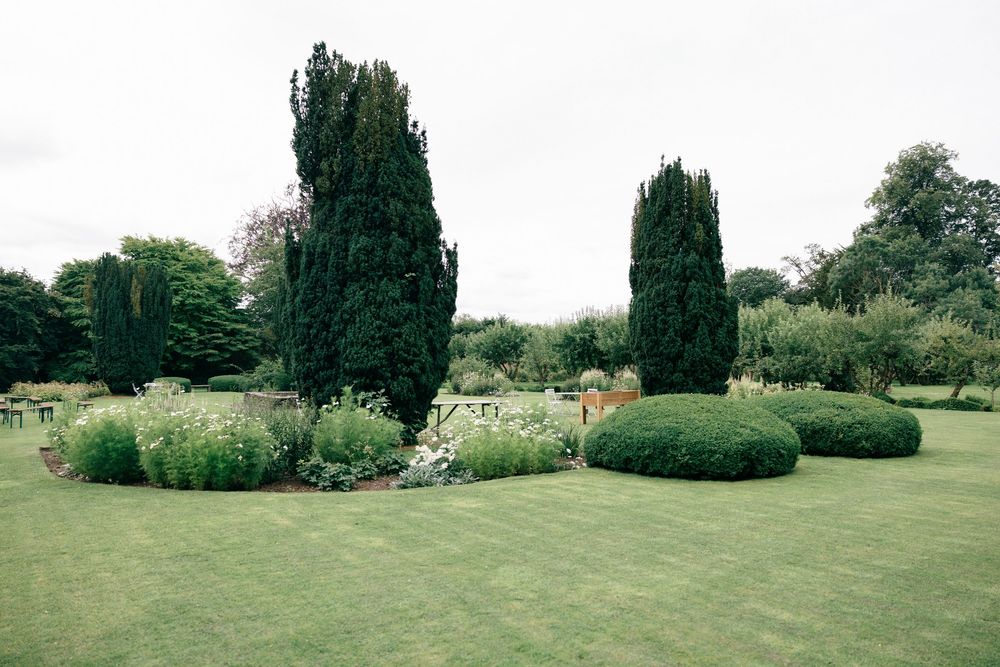 Lush green garden with tall cypress trees, manicured shrubs, and colorful flower beds on a cloudy day.