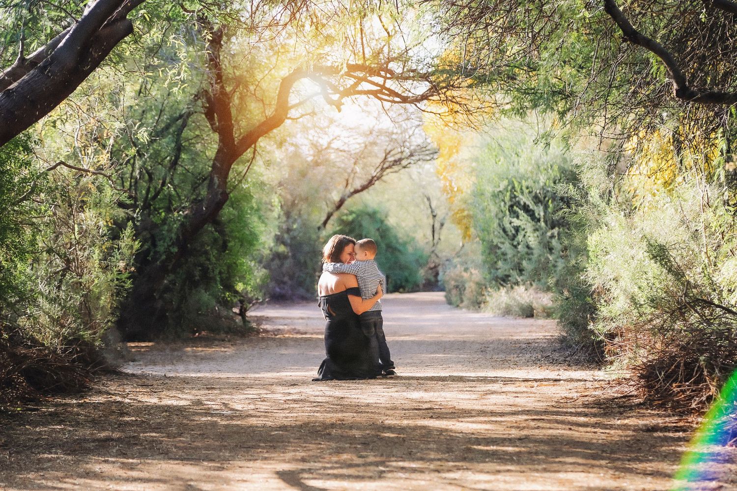 Mother and child embrace on a sunlit dirt path surrounded by green trees, with warm golden light filtering through the leaves.
