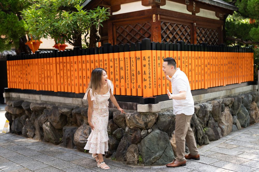 Couple in white outfits standing and smiling beside a stone wall and orange wooden fence at a traditional Japanese temple.