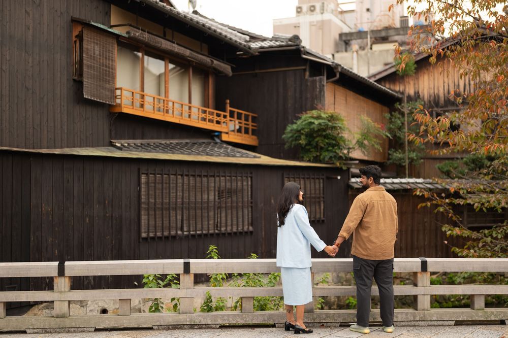 Couple holding hands walk past traditional dark wooden Japanese townhouses with balconies on a quiet historic street in autumn.