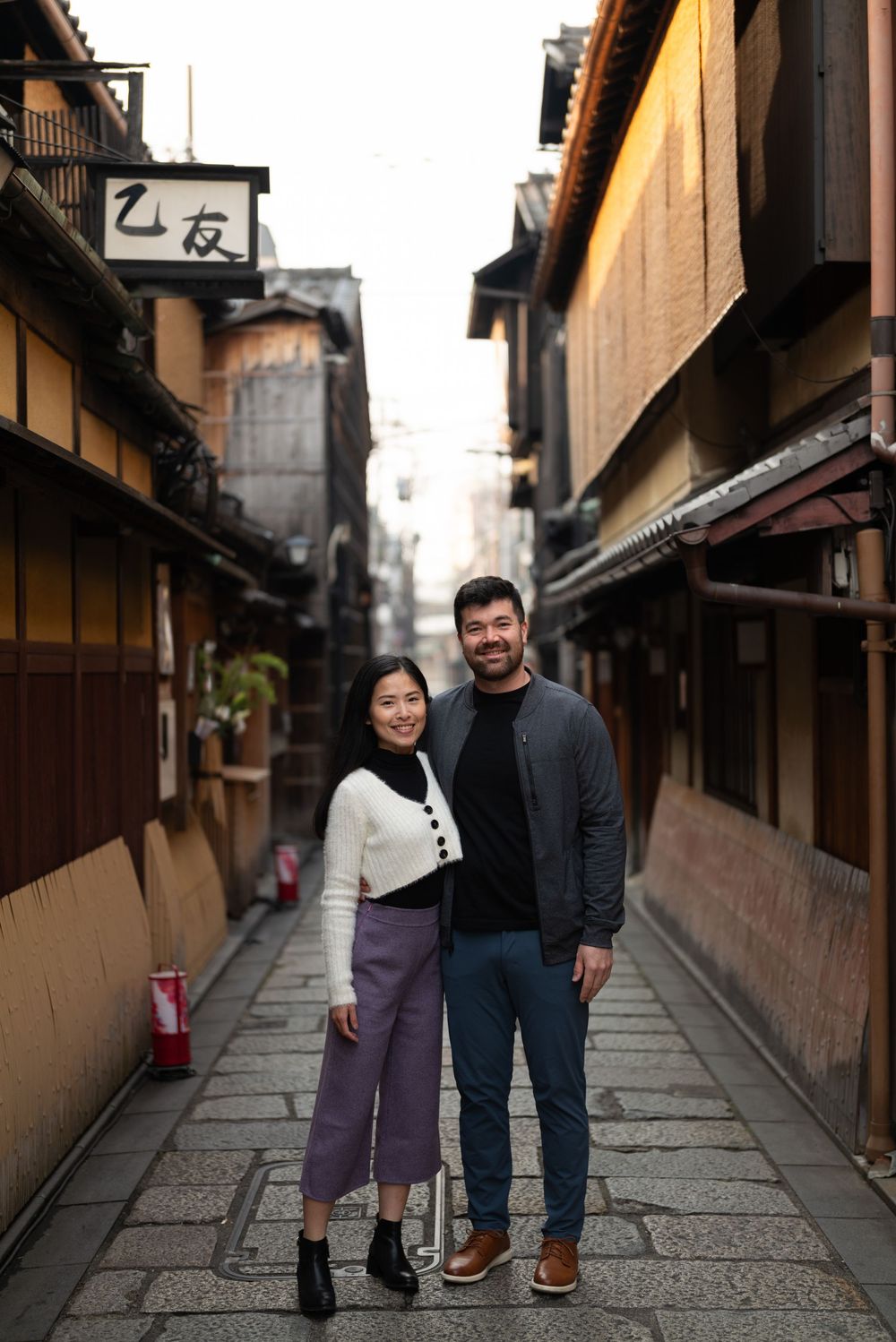 Smiling couple posing together on a stone-paved alley lined with traditional wooden buildings in a historic Kyoto district.