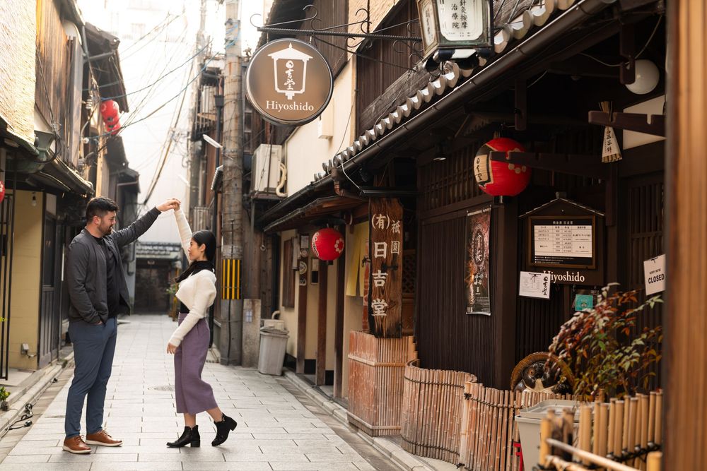 Couple poses playfully on a quiet traditional Kyoto street lined with wooden machiya houses, red paper lanterns and hanging shop signs.