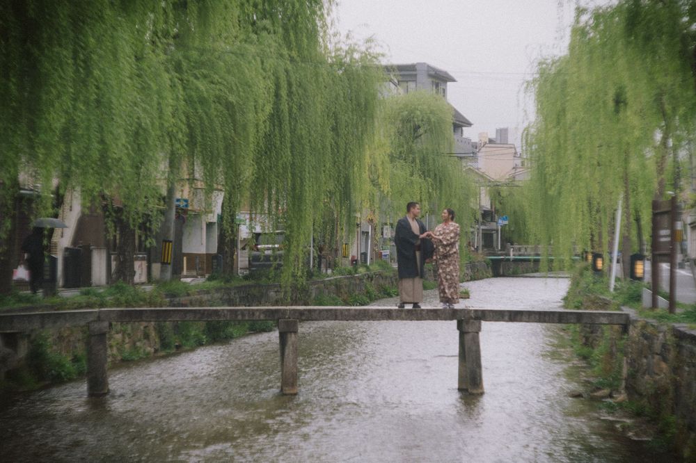 Two people in traditional kimono stand on a narrow stone bridge over a calm canal lined with weeping willow trees.
