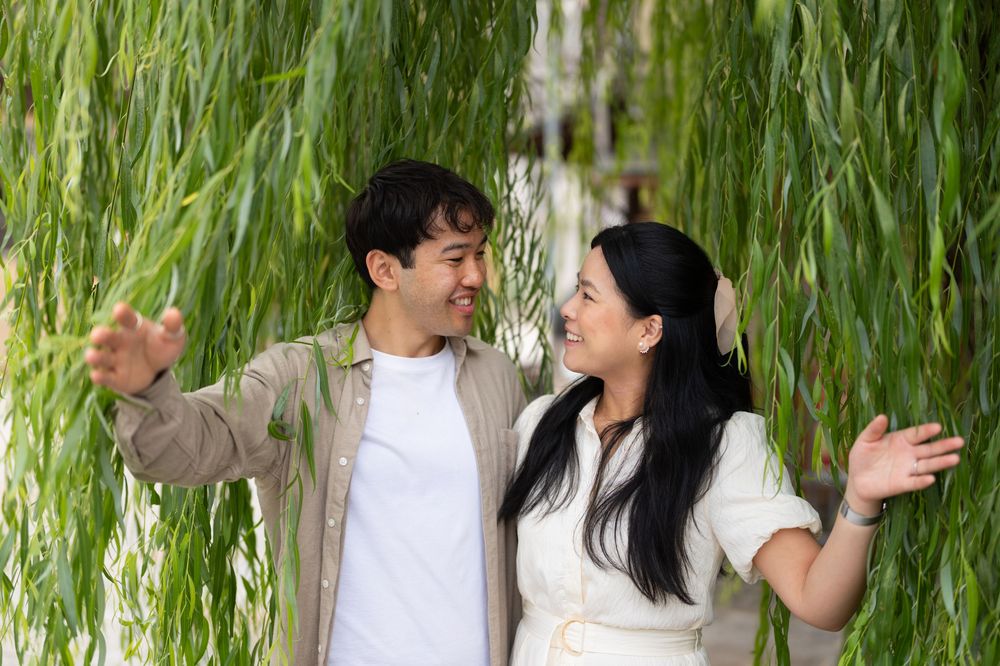 A smiling young man and woman stand beneath weeping willow branches, arms outstretched, enjoying a happy moment together outdoors.
