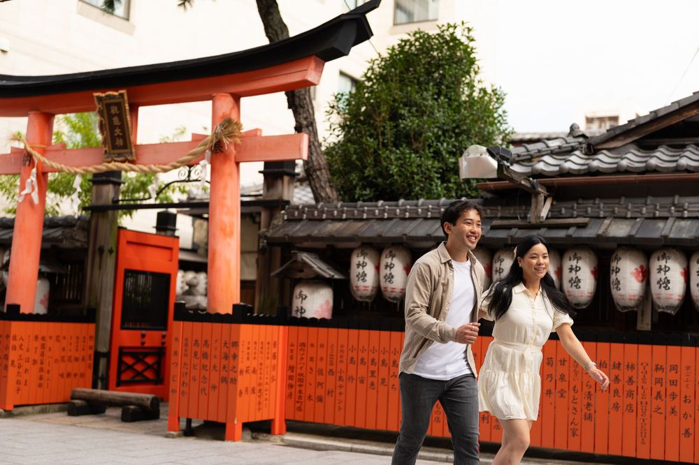 Smiling couple walks hand in hand past a vermilion torii gate and traditional wooden shrine with paper lanterns in Kyoto, Japan.