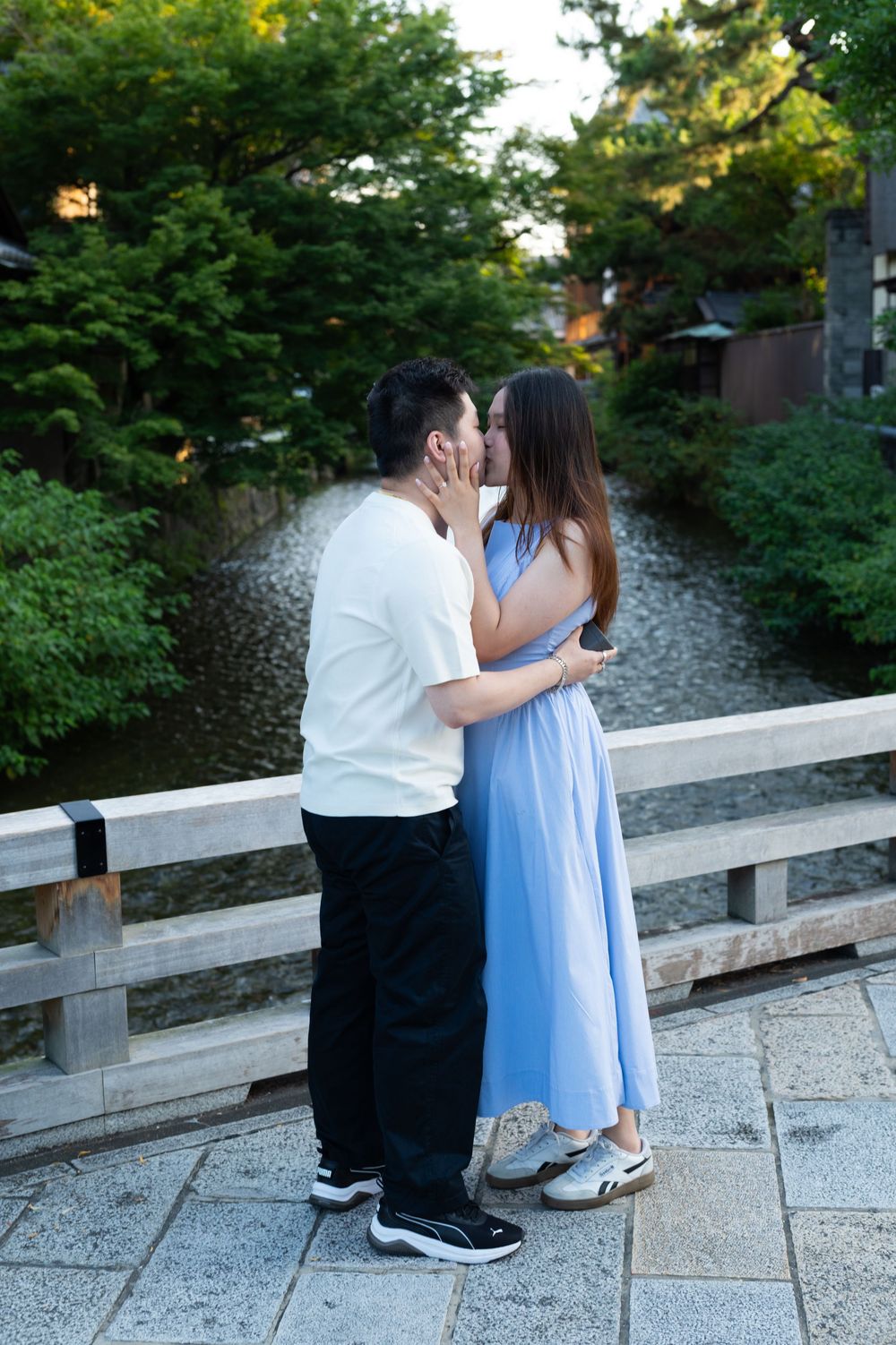 Couple in casual clothes kissing on a wooden bridge over a calm stream, surrounded by lush green trees on a stone-paved path.