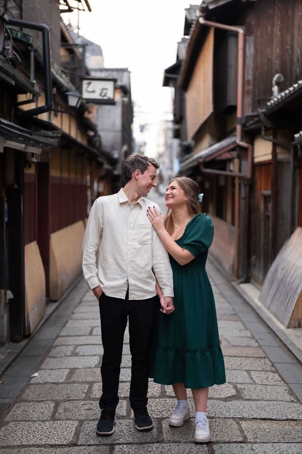Smiling couple holding hands and gazing at each other on a stone-paved traditional Japanese street lined with wooden buildings in Kyoto.