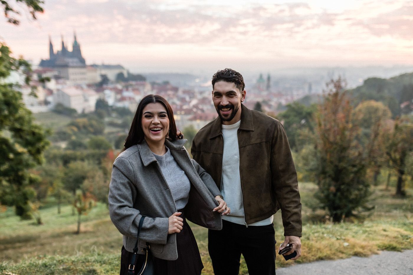 Smiling couple sharing a sweet moment on a hillside at sunset with a hazy historic city skyline and castle spires in the background.