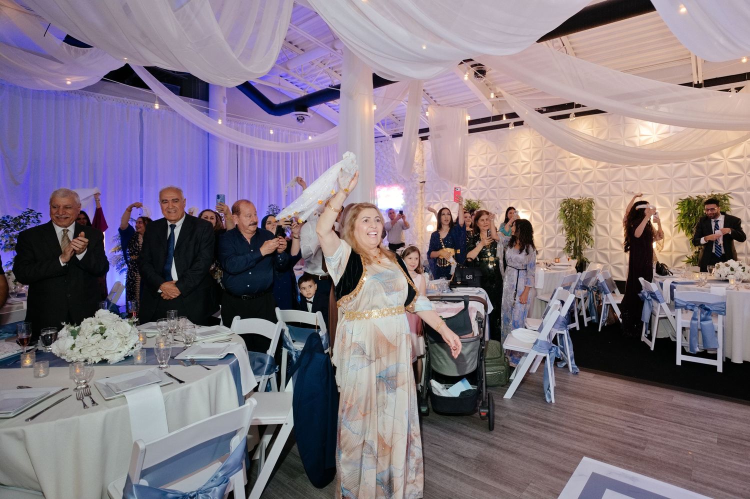 Woman in a floral dress holds a white cloth overhead at a wedding reception while guests in formal wear watch in a draped banquet hall.