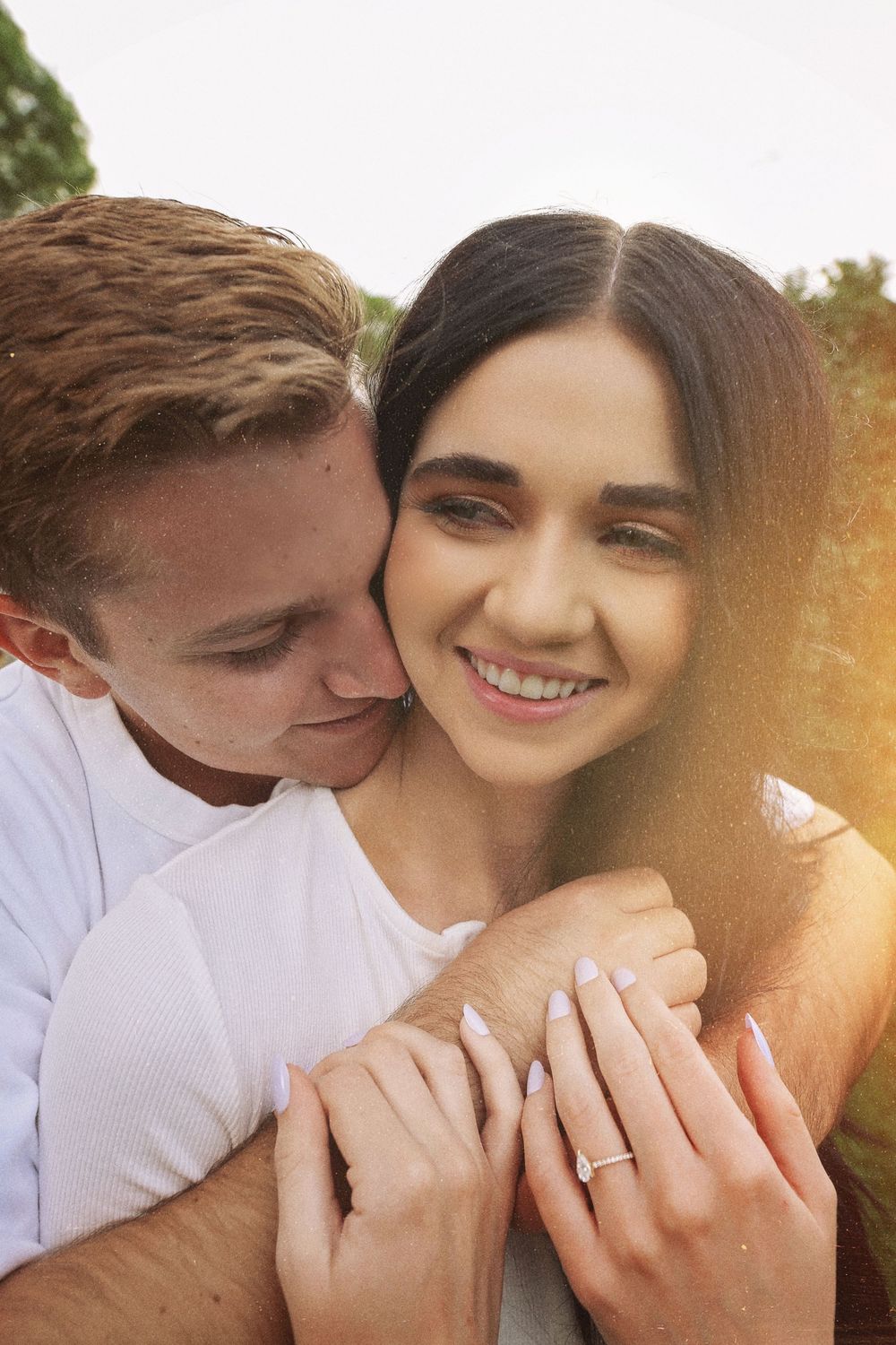 A smiling woman with a diamond ring is hugged from behind by a man in warm, glowing sunlight.