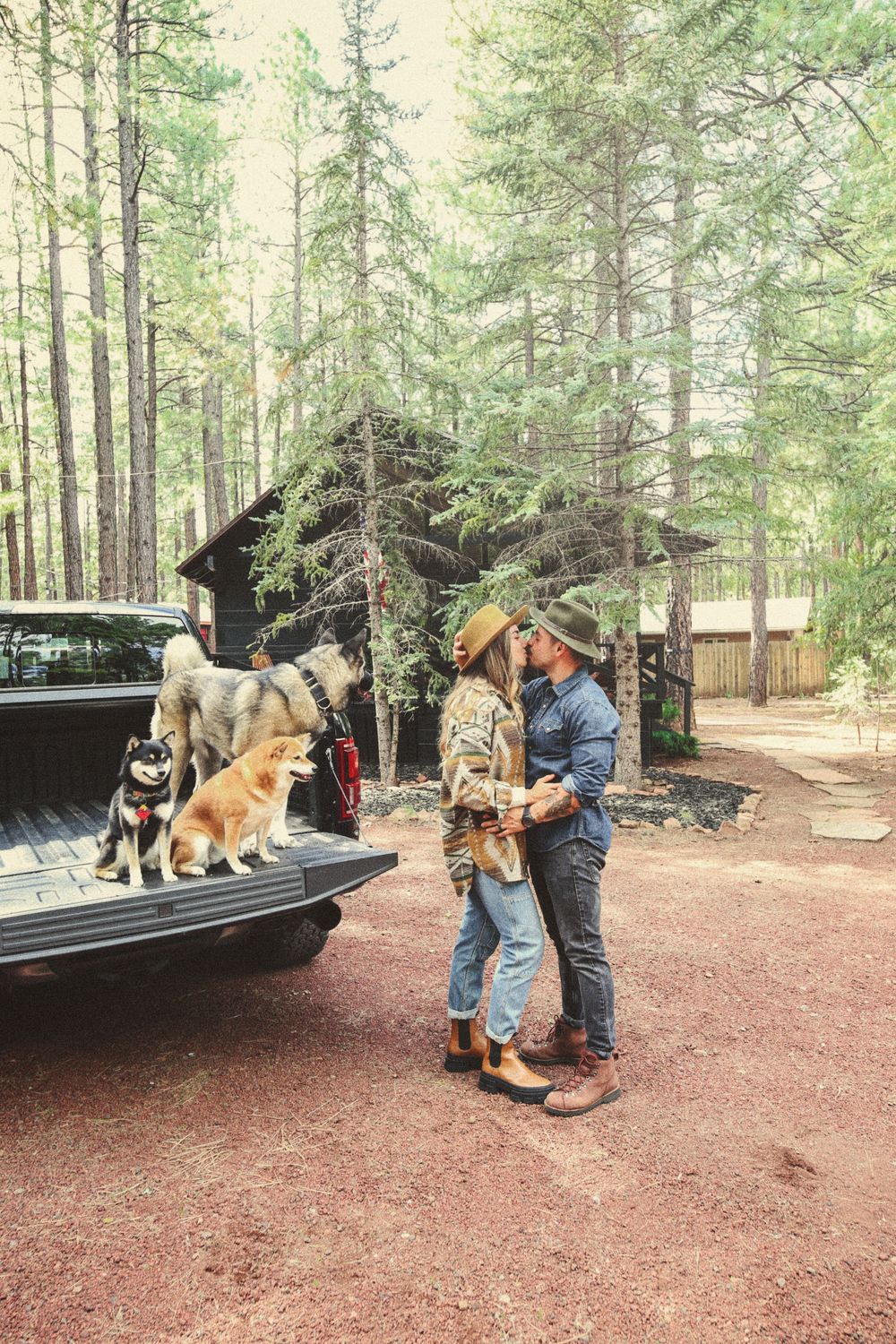 Couple in hats embracing near a pickup truck with three dogs in the bed, beside a rustic cabin in a pine forest.
