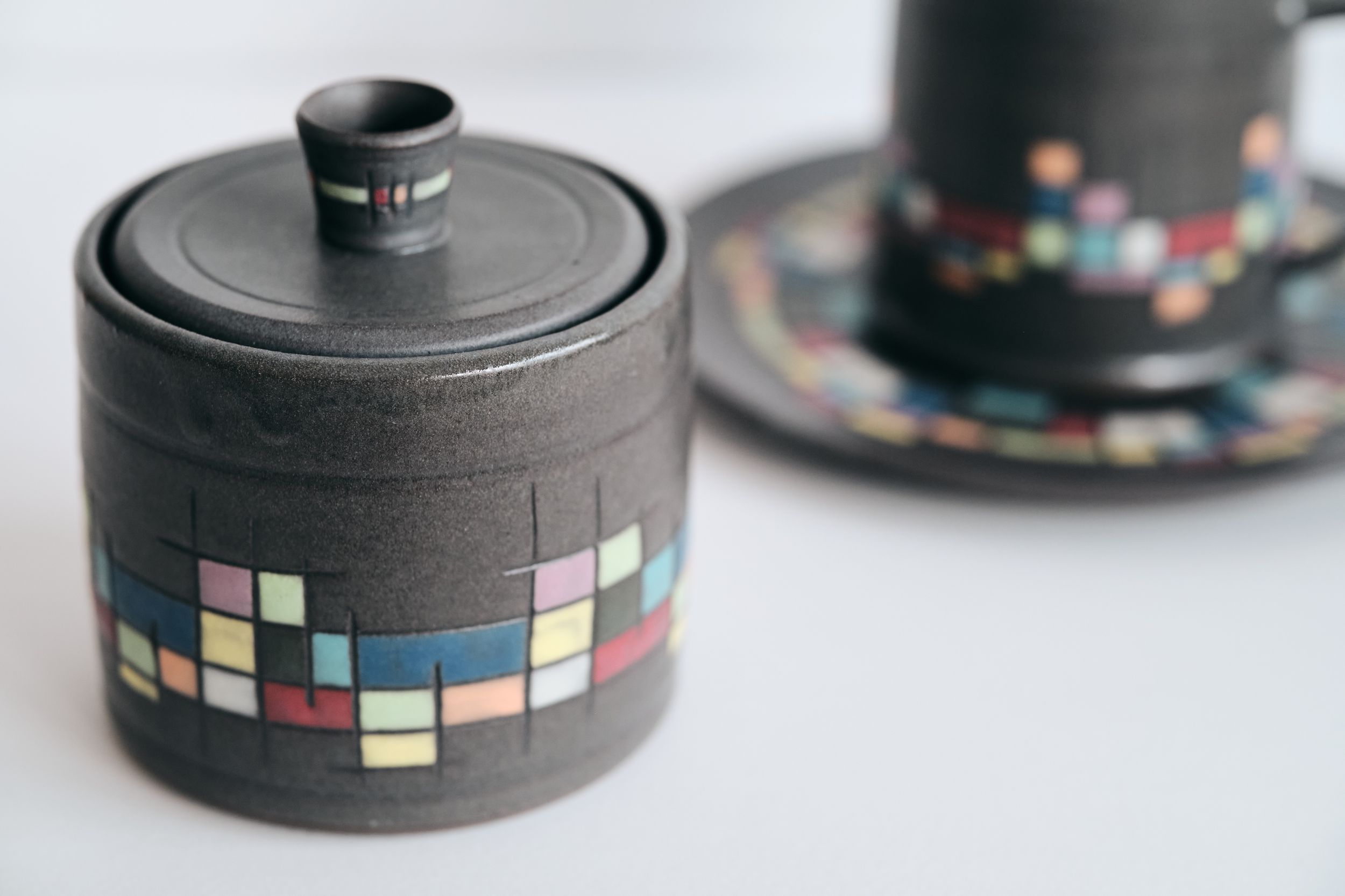 Handmade dark gray ceramic sugar bowl with lid and matching cup and saucer, decorated with colorful mosaic-style tile pattern bands.