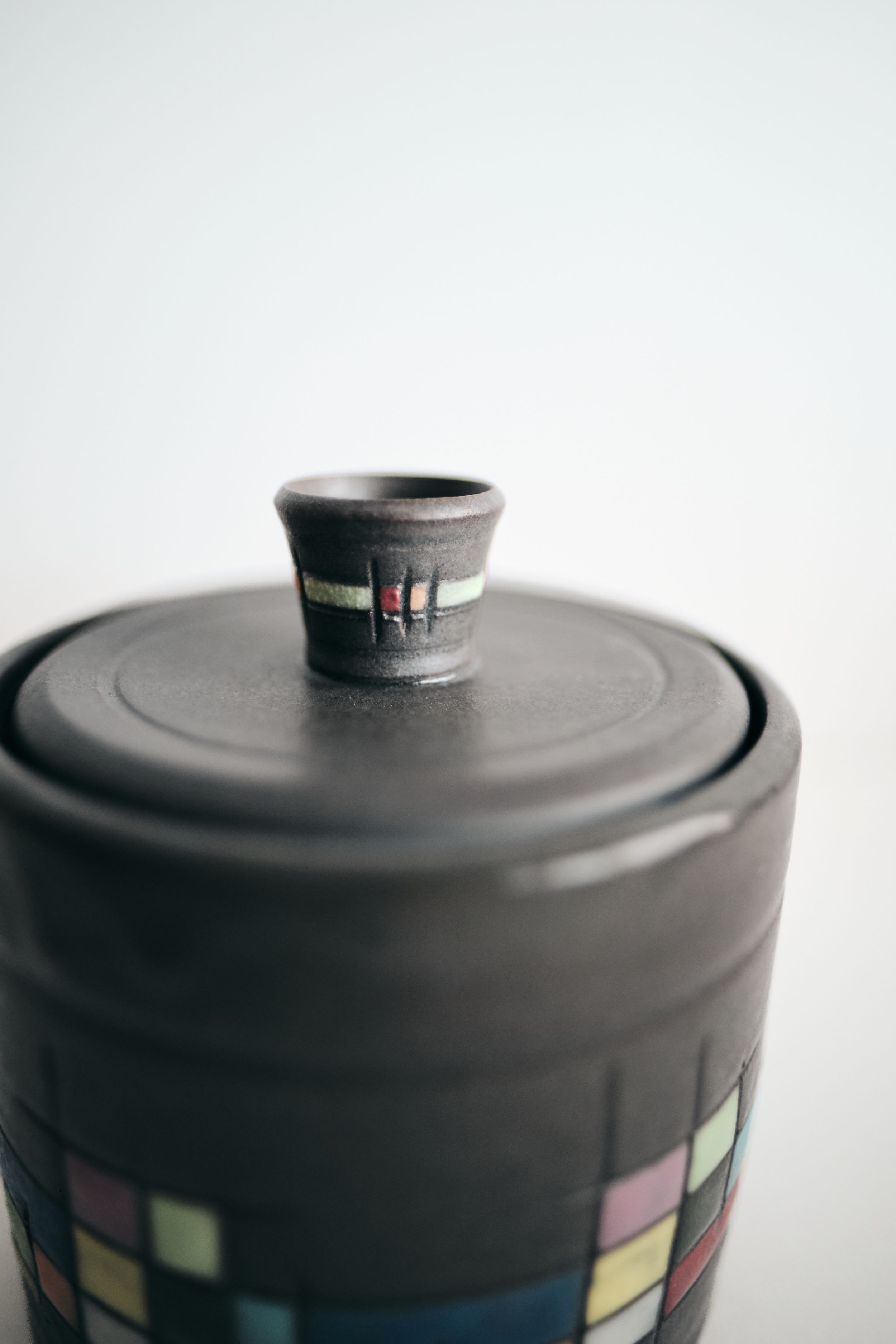 Close-up of a dark handmade ceramic lidded jar with a small knob handle and colorful geometric tile pattern along the base.