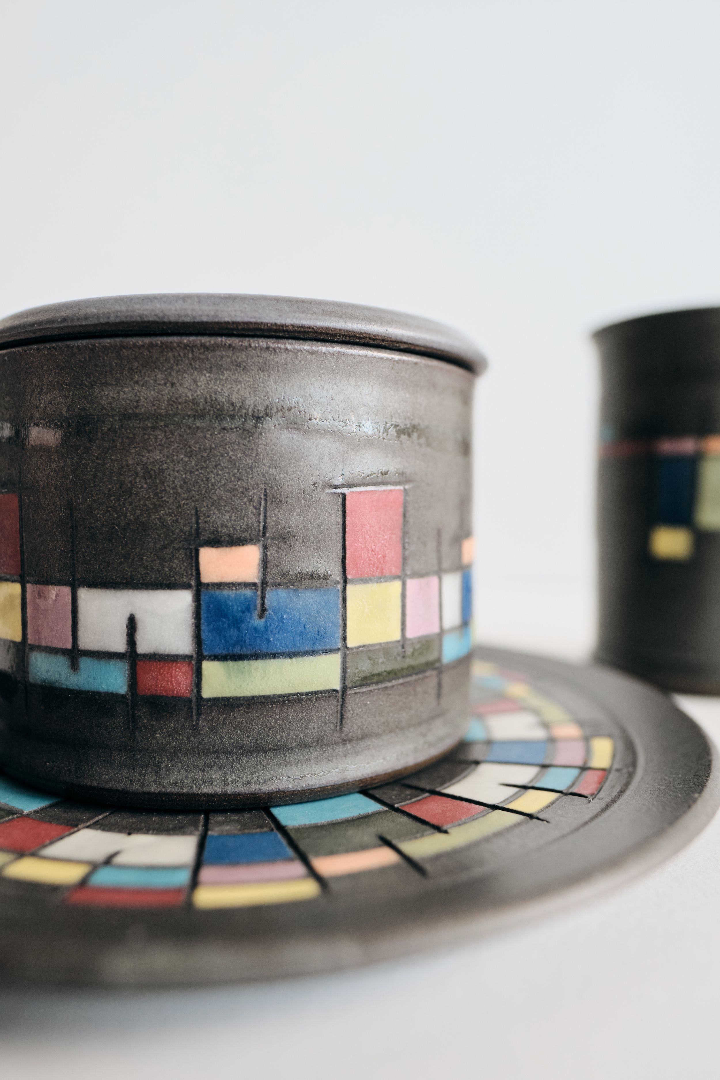 Handmade dark ceramic lidded jar on matching saucer with colorful geometric mosaic band in blue, pink, yellow and red.