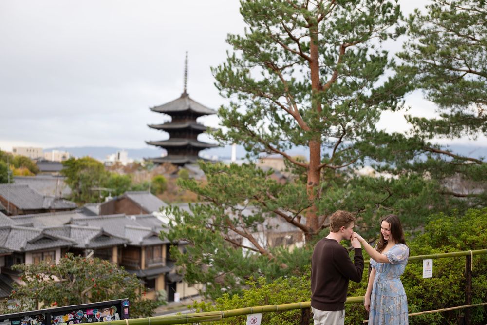 Couple standing at a scenic overlook in Kyoto, with a traditional pagoda, pine trees and historic wooden rooftops in the background.